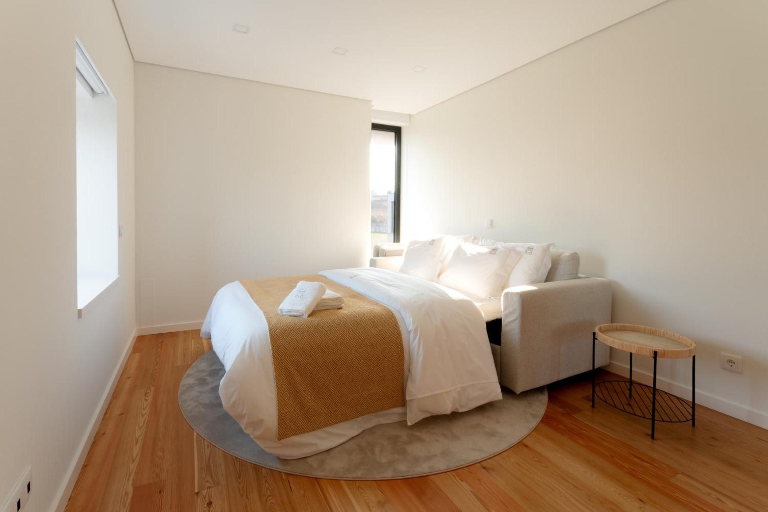 Un dormitorio minimalista con una cama, una mesita redonda pequeña y un ventanal que deja entrar luz natural.