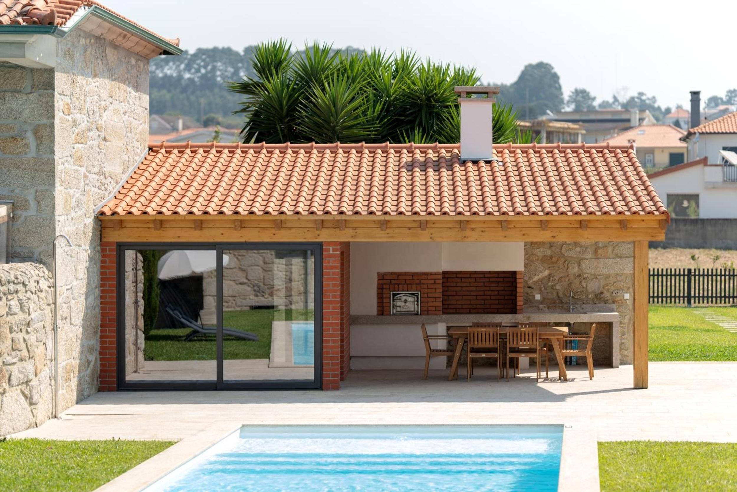 Una casa moderna con piscina, un porche cubierto con mesa y sillas, y un muro de piedra.