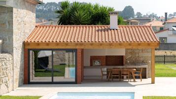 Una casa moderna con piscina, un porche cubierto con mesa y sillas, y un muro de piedra.