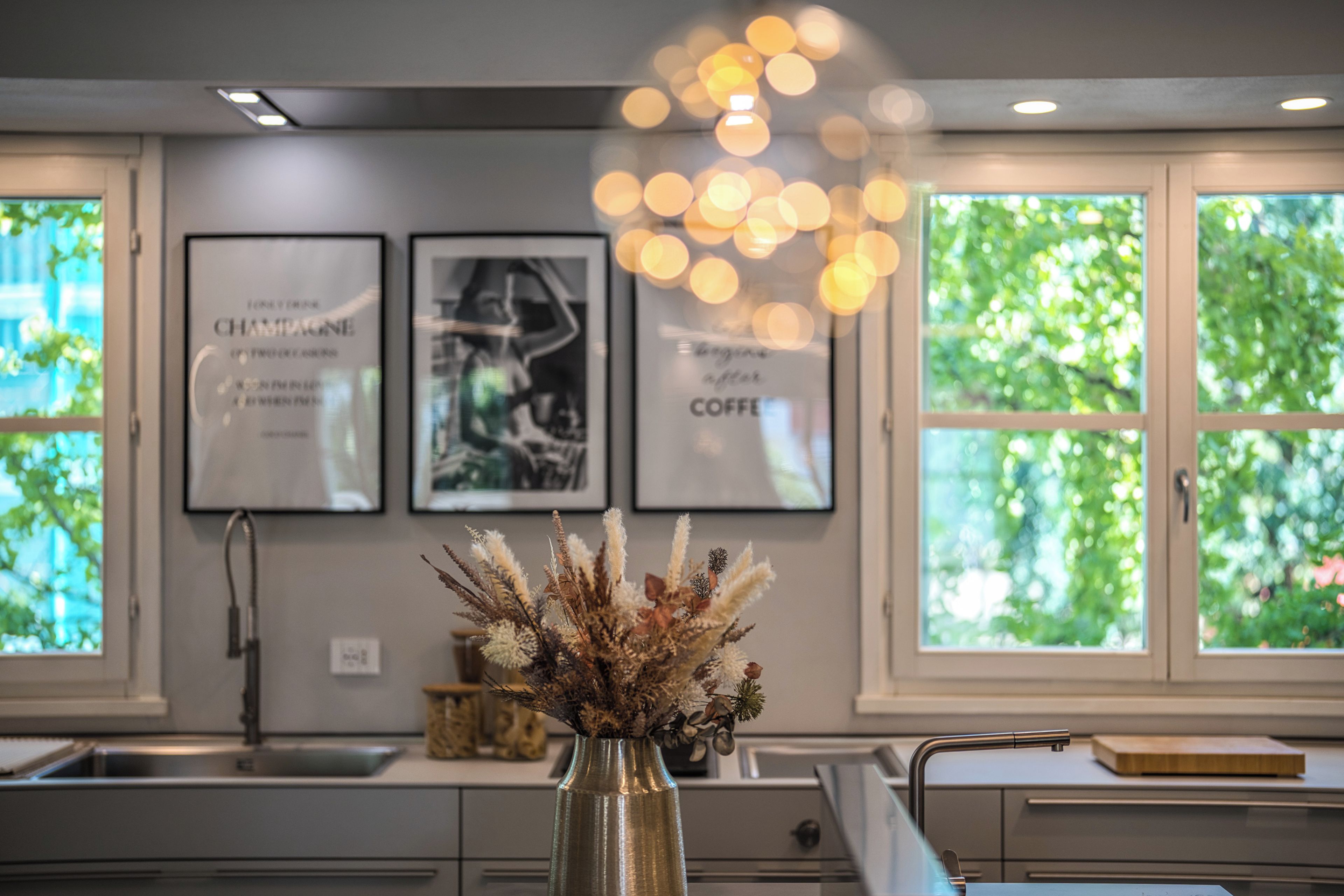 A kitchen with a vase of dried plants on the counter, a sink, and framed pictures on the wall.