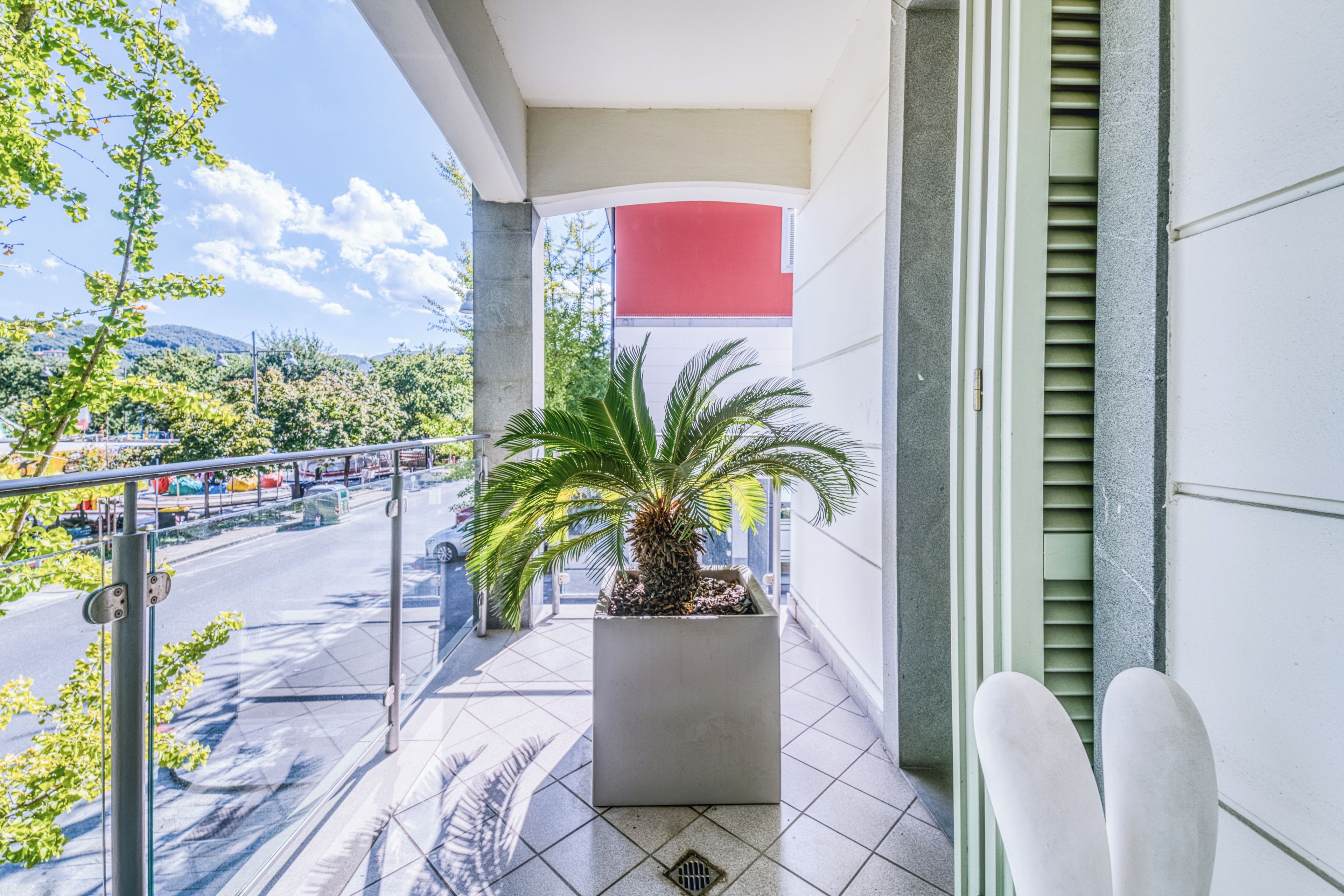Un balcón con una palmera en maceta, vista de una carretera con vehículos y un edificio con una sección roja.