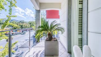 Un balcón con una palmera en maceta, vista de una carretera con vehículos y un edificio con una sección roja.
