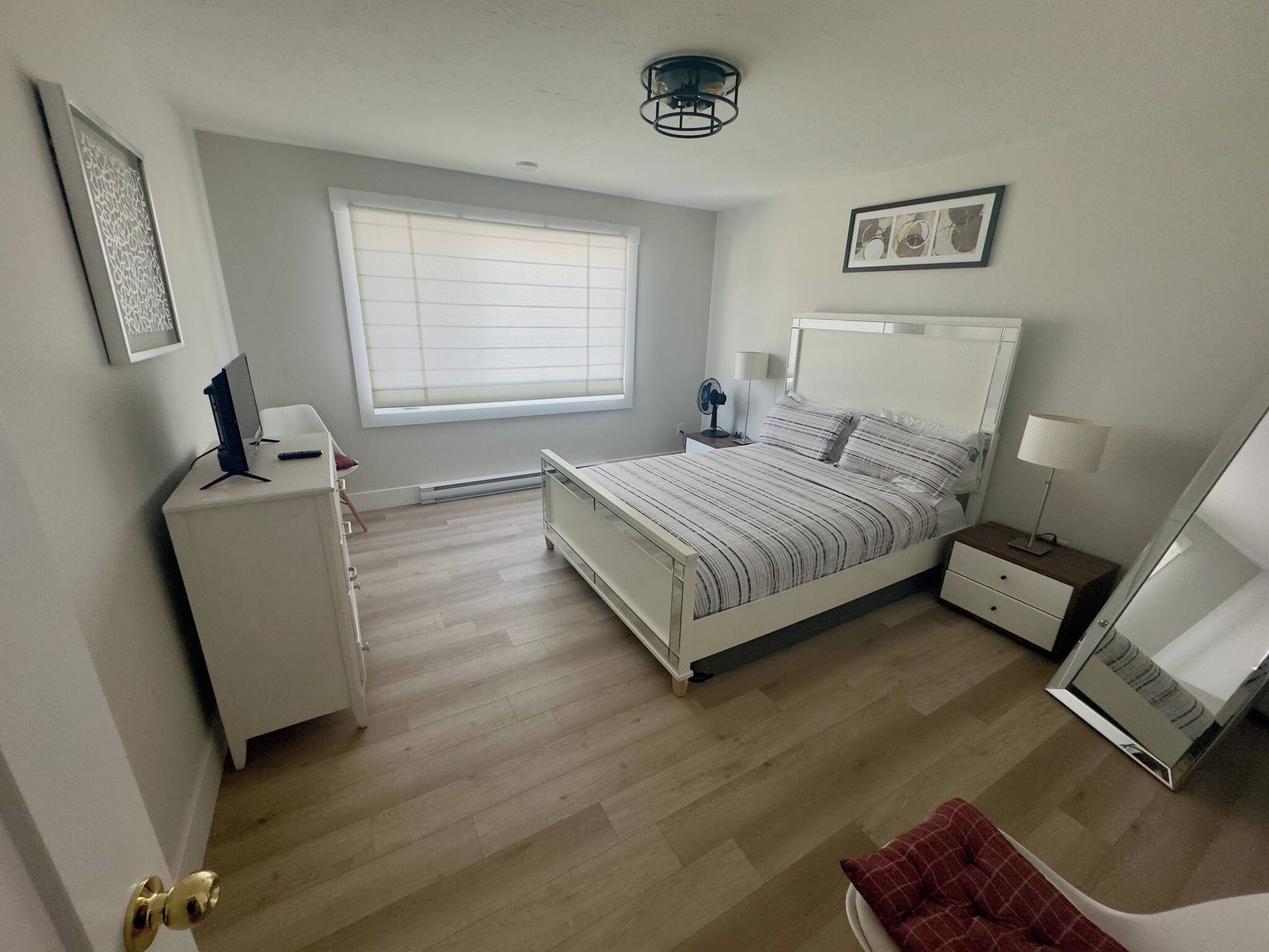 A bedroom with a bed, bedside tables, a desk, a chair, a window with blinds, and a ceiling light fixture.