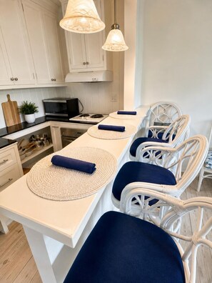 A kitchen with white cabinets, a black microwave, and a stove. There are two hanging pendant lights and a white island with blue cushions and woven placemats. - Luxury Loft |Sauna, Hot Tub & Peaceful BalconyView (Houston)