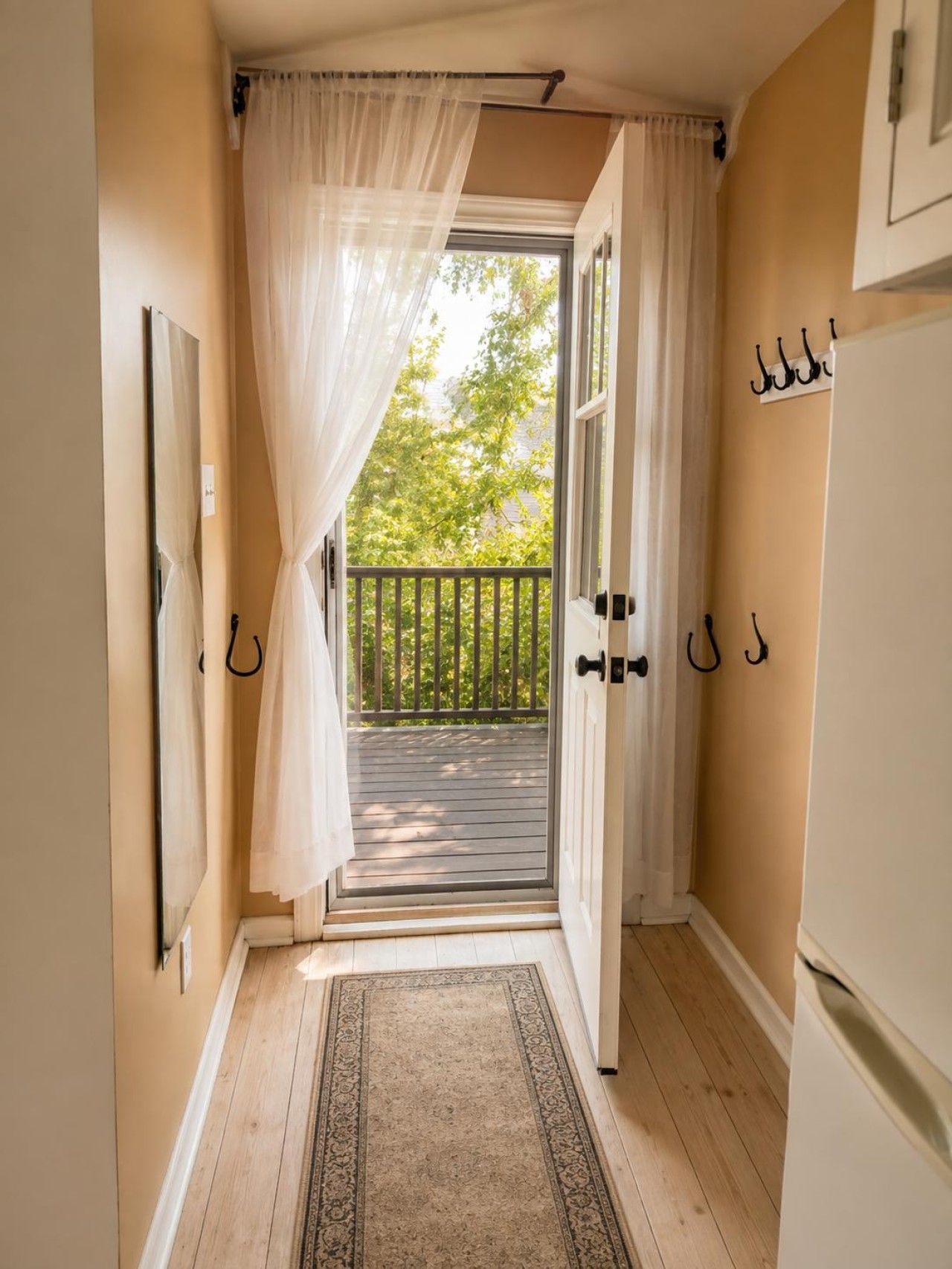 A narrow hallway with a wooden floor, a white door leading to a balcony, and a white curtain partially drawn.