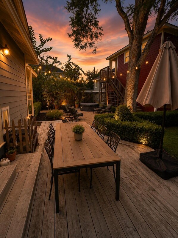 Een buitenterras met een houten tafel en stoelen, omringd door een houten vlonder, een jacuzzi en een trap naar een rood gebouw.