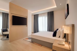 A modern hotel room with a large bed, a flat-screen TV mounted on a wooden panel, a bedside table with a lamp, and a chair.