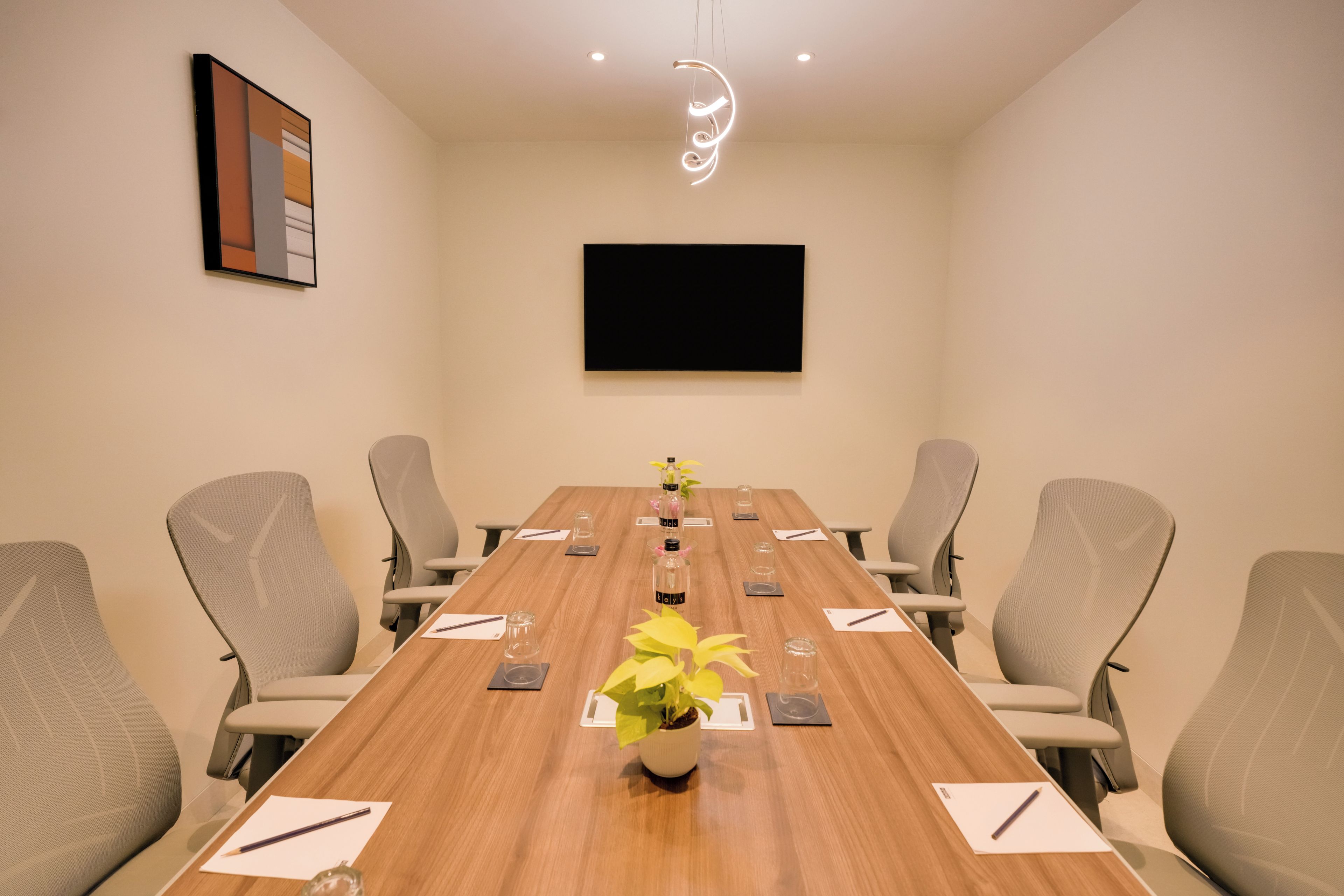 Un salón de conferencias con una larga mesa de madera, seis sillas, un televisor de pantalla plana en la pared y una lámpara decorativa.