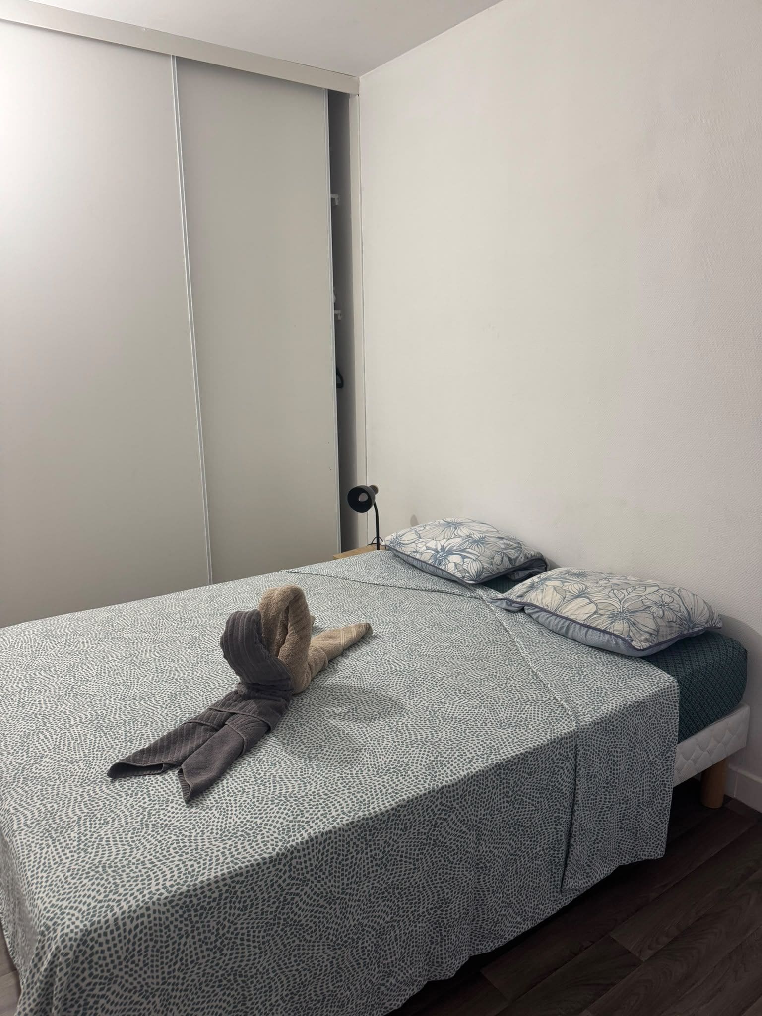 Un lit simple avec une couvre-lit à motifs et des oreillers, une table de chevet avec une lampe et une armoire fermée.