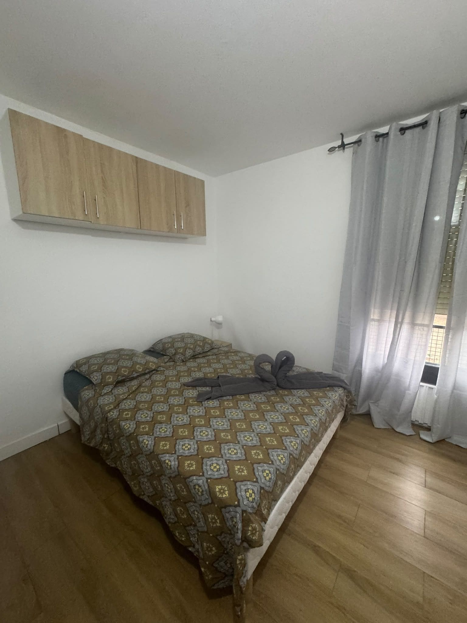 A bedroom with a bed, a wooden wardrobe, a window with curtains, and a light switch on the wall.