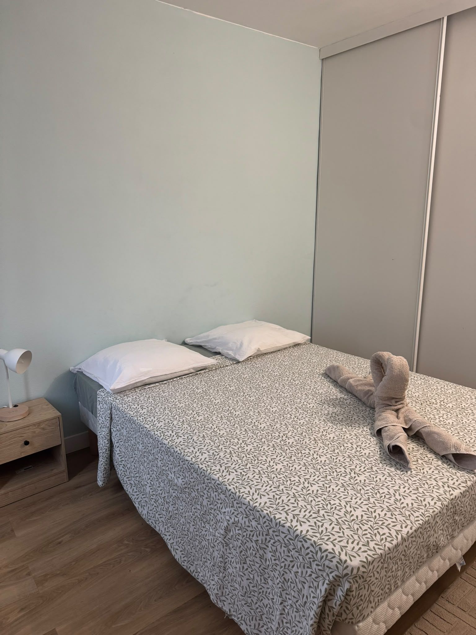 Un lit simple avec une couvre-lit à motifs, une table de chevet avec une lampe et une armoire fixée au mur.