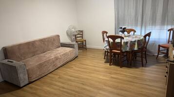 A living room with a sofa, a dining table with chairs, a fan, and a window with curtains.