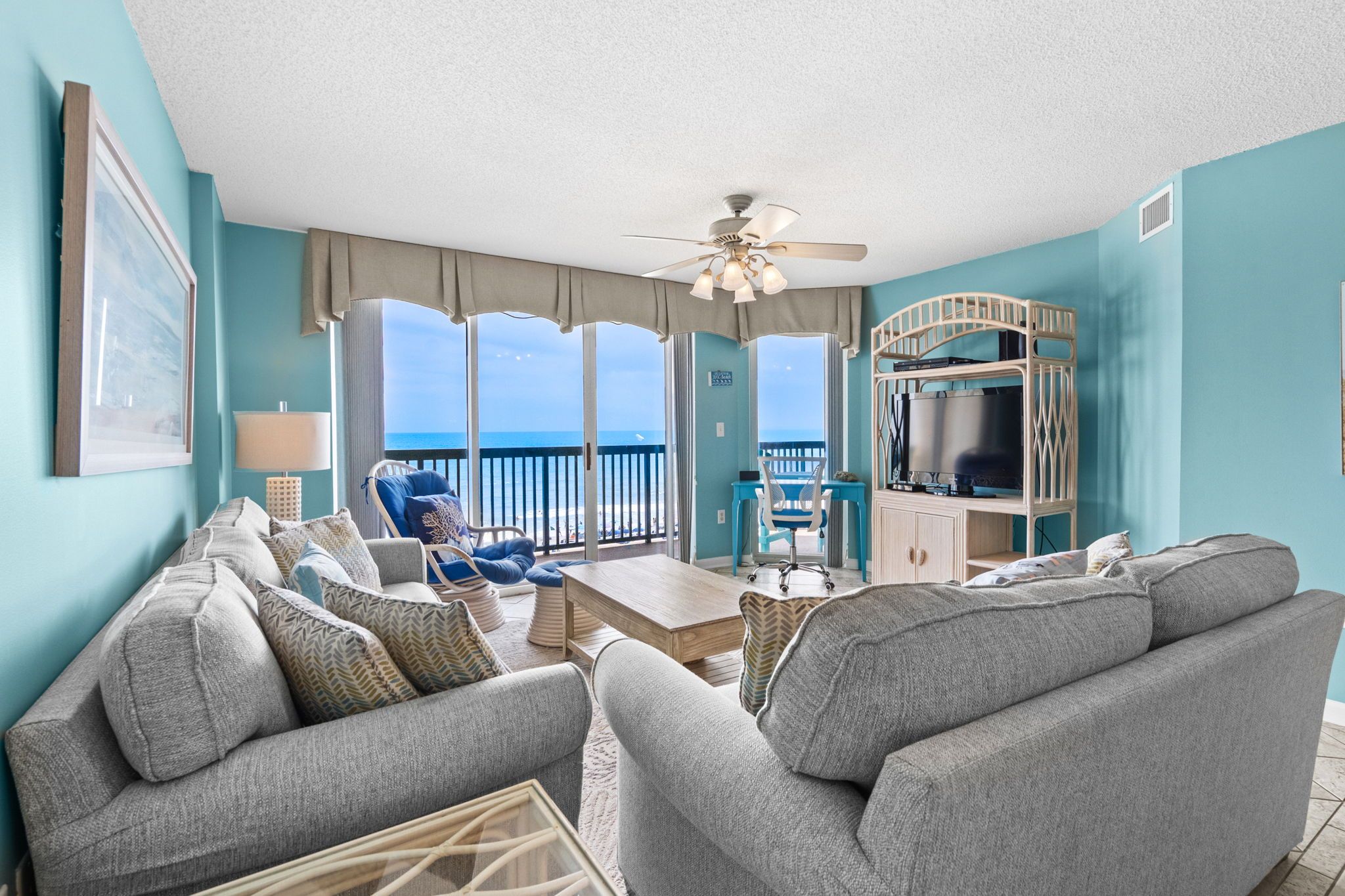 A living room with a view of the ocean, a ceiling fan, and a TV on a wooden stand.