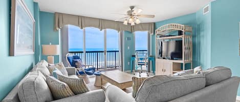 A living room with a view of the ocean, a ceiling fan, and a TV on a wooden stand.
