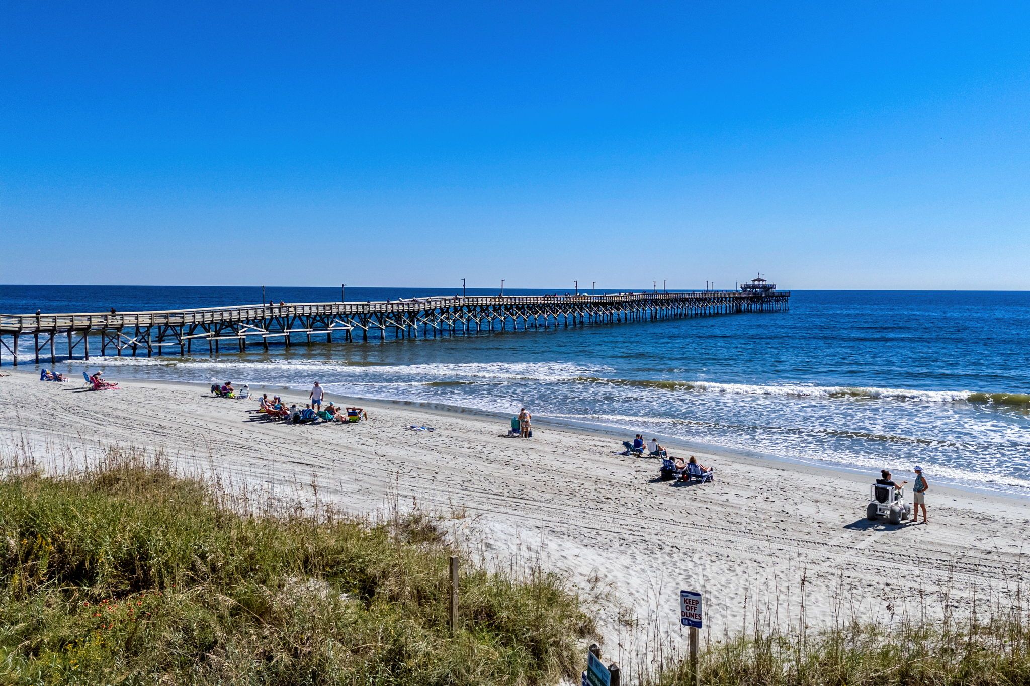 A beach with a pier extending into the ocean, people relaxing on the sand, and a sign indicating 'KEEP OFF DUNES'.
