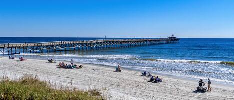 A beach with a pier extending into the ocean, people relaxing on the sand, and a sign indicating 'KEEP OFF DUNES'.