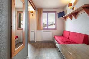 A room with a wooden table, a red sofa, a window with curtains, and a wall-mounted lamp. - Apartment near Ski Lifts (Tignes)