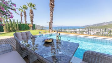 Un área de piscina exterior con una mesa lista para comer, rodeada de muebles de mimbre, palmeras y vistas al mar.