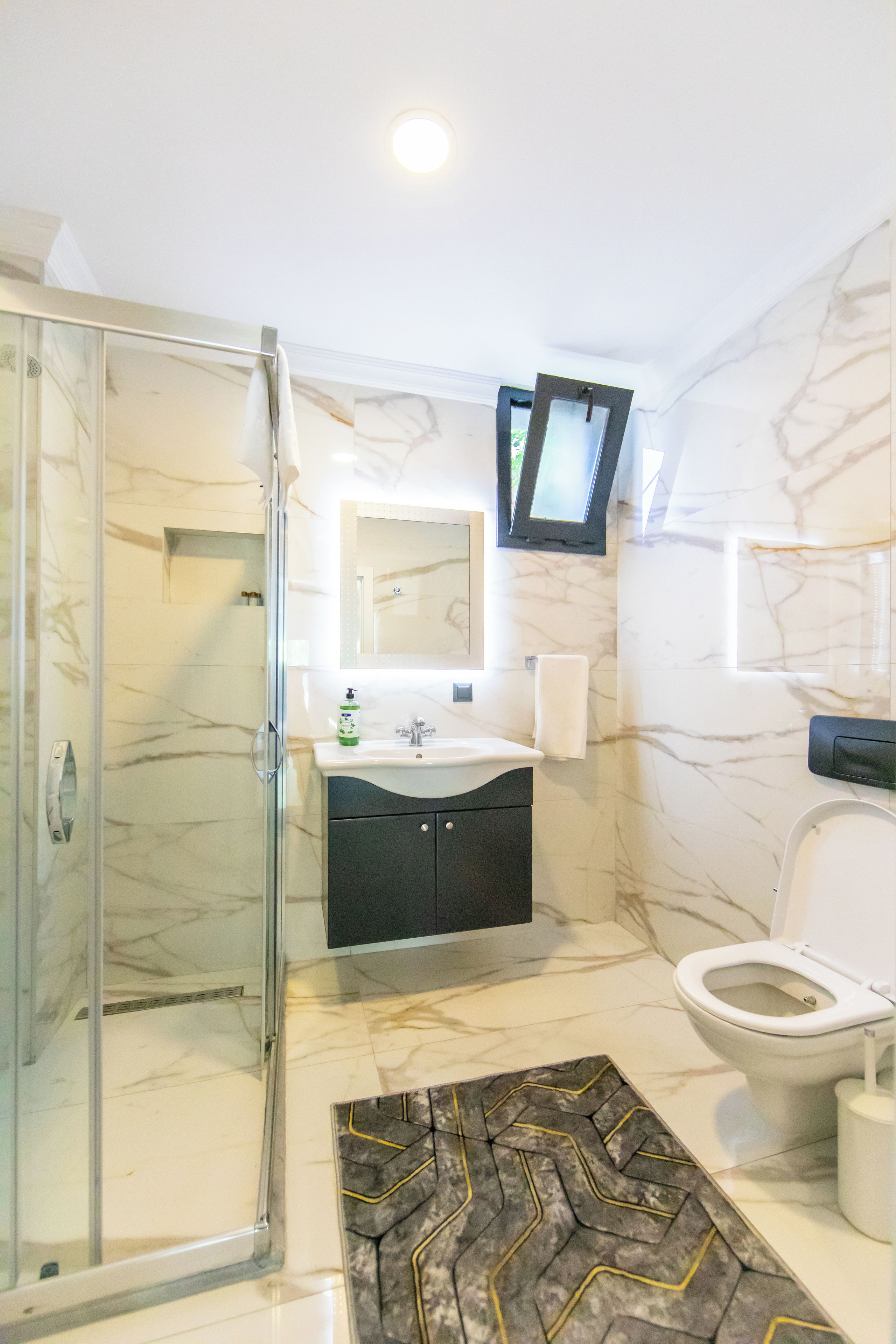 Ein Badezimmer mit Duschkabine aus Glas, Waschbecken mit Spiegel darüber, Toilette und einem an der Wand montierten Fernseher.
