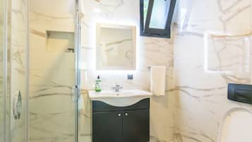 Ein Badezimmer mit Duschkabine aus Glas, Waschbecken mit Spiegel darüber, Toilette und einem an der Wand montierten Fernseher.