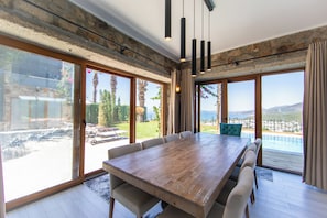 Un comedor amplio con una gran mesa de madera y varias sillas, ventanas grandes con vista a una piscina y paisaje, y una pared de piedra.
