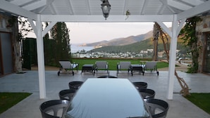 Un espace extérieur abrité, aménagé pour les repas, avec une table et des chaises, une vue sur la mer et les collines en arrière-plan, et une structure en forme de pergola.