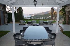 A covered outdoor dining area with a table and chairs, a view of the sea and hills in the background, and a pergola structure. - Mira villa 2 (Torba)