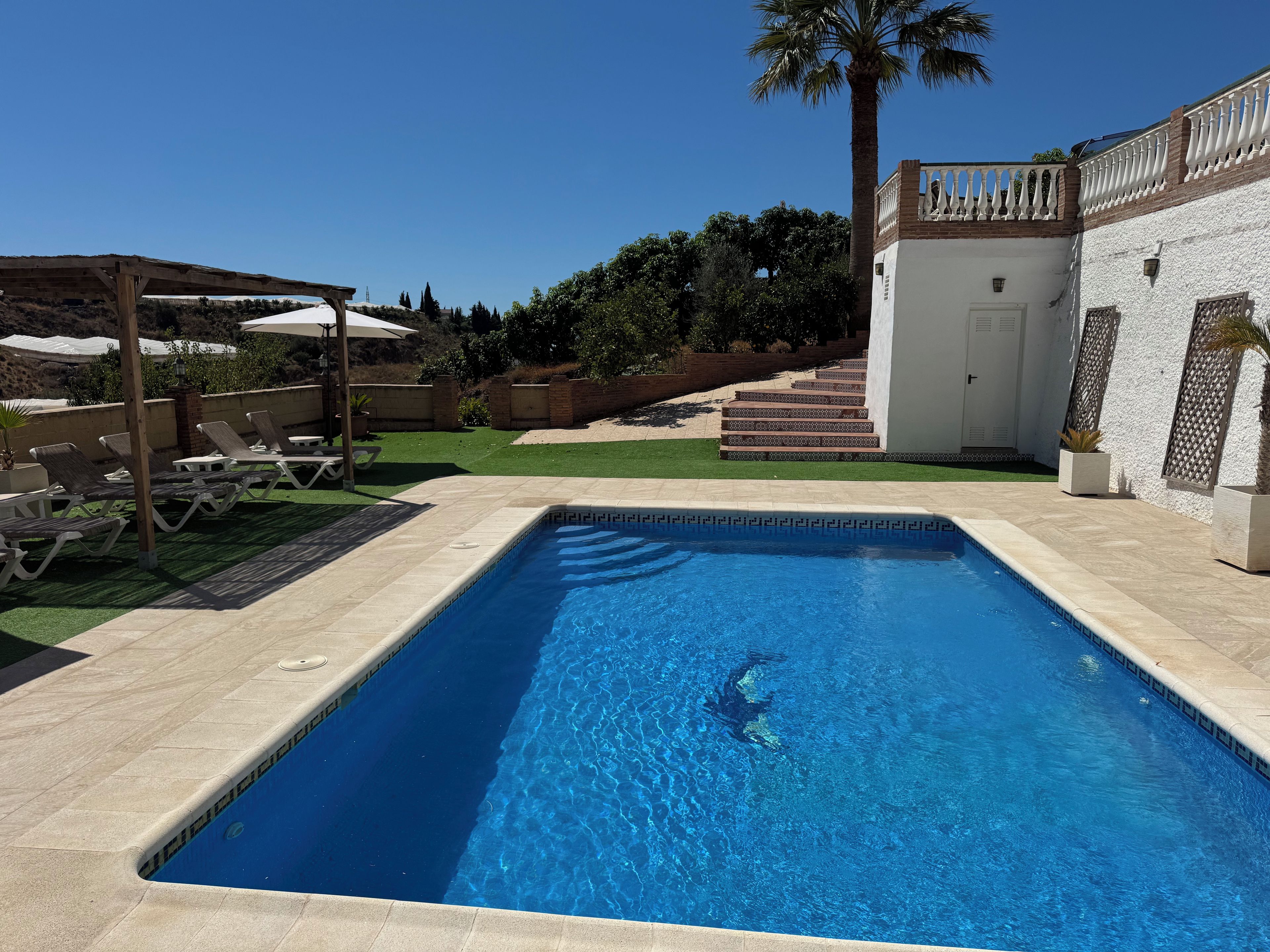 A rectangular swimming pool with clear blue water, surrounded by a tiled deck, a covered seating area with umbrellas, and a white building with a balcony.