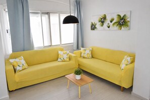 Ein gelbes Sofa mit passenden Kissen, ein kleiner Holztisch mit einer Pflanze und eine Wand mit einem floralen Gemälde.