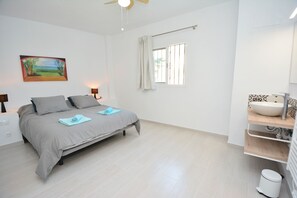 Un dormitorio con cama, un cuadro, una ventana con cortinas, una lámpara de noche y un baño con lavamanos e inodoro.