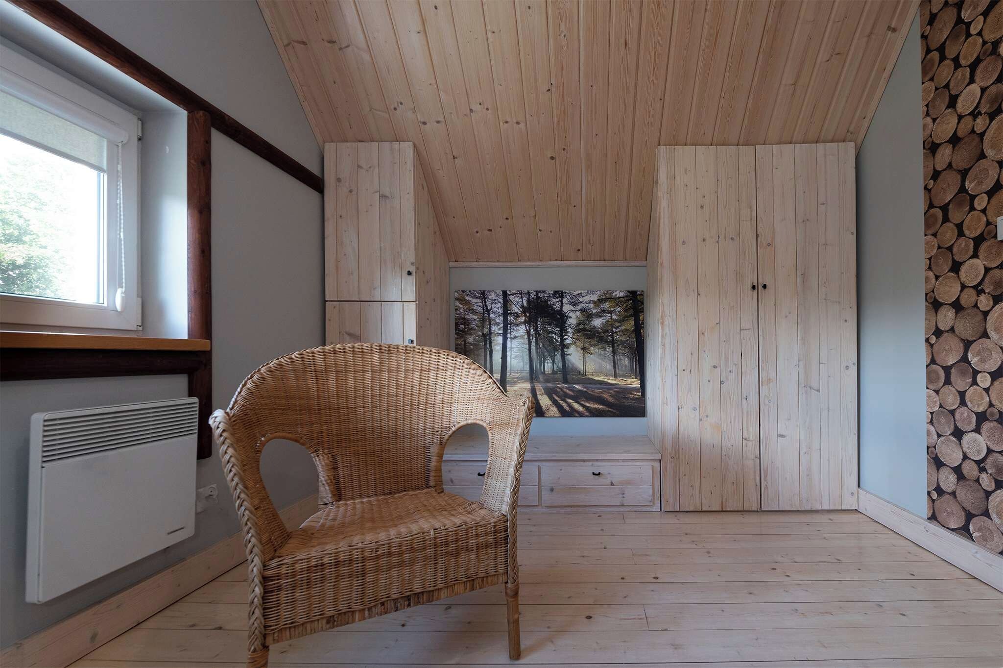 Une pièce avec un fauteuil en osier, une grande fenêtre, des murs en bois et une vue sur une forêt, visible à travers un tableau.