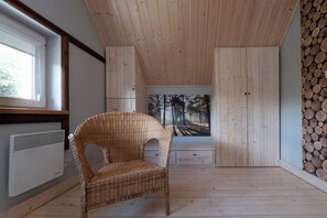 Une pièce avec un fauteuil en osier, une grande fenêtre, des murs en bois et une vue sur une forêt, visible à travers un tableau.