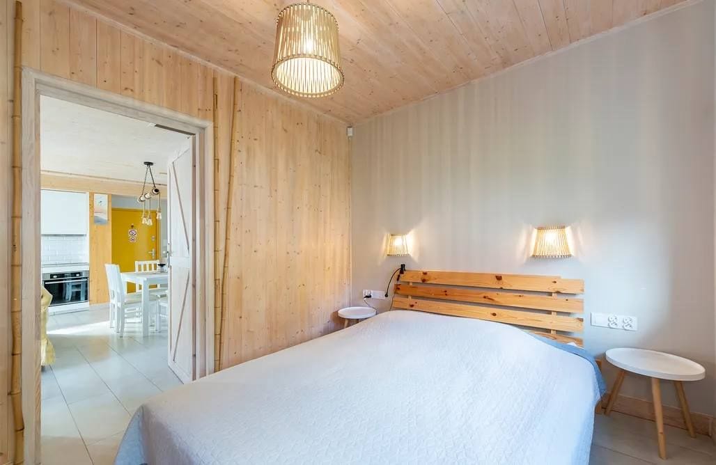 A bedroom with a wooden headboard, a bed, bedside tables, and a door leading to a dining area.