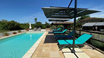 Piscina com espreguiçadeiras e guarda-sóis, ladeada por um terraço de pedra.
