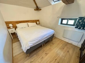 Ein Schlafzimmer mit einem hölzernen Bett, weißer Bettwäsche, einem Nachttisch mit Lampe, einem kleinen Fenster und einem Wandheizgerät.