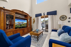 Ein Wohnzimmer mit Fernseher, blauen Sofas, einem Holzschrank und Blick durch eine Glastür auf einen Außenbereich.