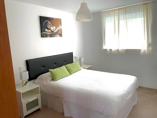 A bedroom with a bed, bedside tables, a nightstand, a lamp, a window with curtains, and a wall-mounted picture.