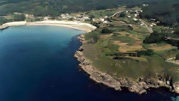 Eine Luftaufnahme einer Küstenlandschaft mit Strand, sanften Hügeln und verstreuten Gebäuden.
