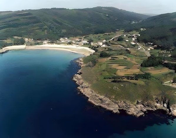 Vista aèria d'una zona costanera amb una platja, turons suaus i edificis dispersos.