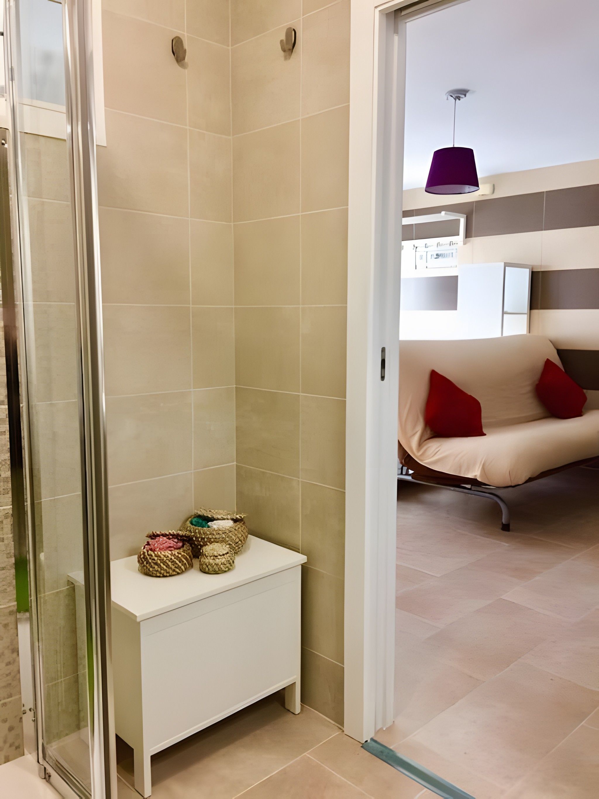 Un baño moderno con baldosas color crema, mampara de vidrio y una mesita de noche blanca con cestas decorativas.