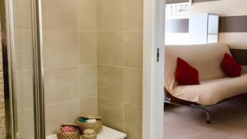 Un baño moderno con baldosas color crema, mampara de vidrio y una mesita de noche blanca con cestas decorativas.
