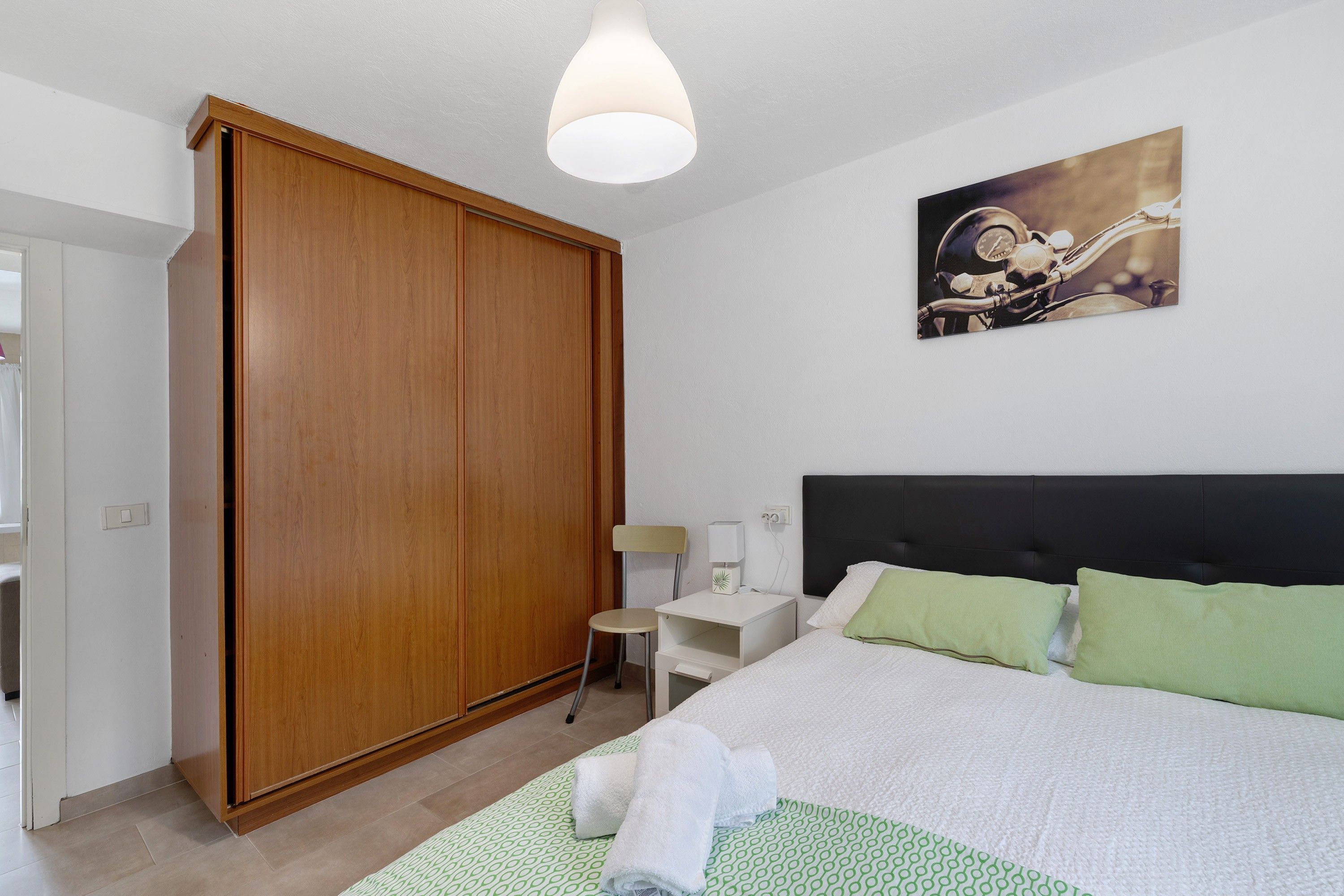 Un dormitorio con cama, armario, mesita de noche, lámpara y un cuadro colgado en la pared.