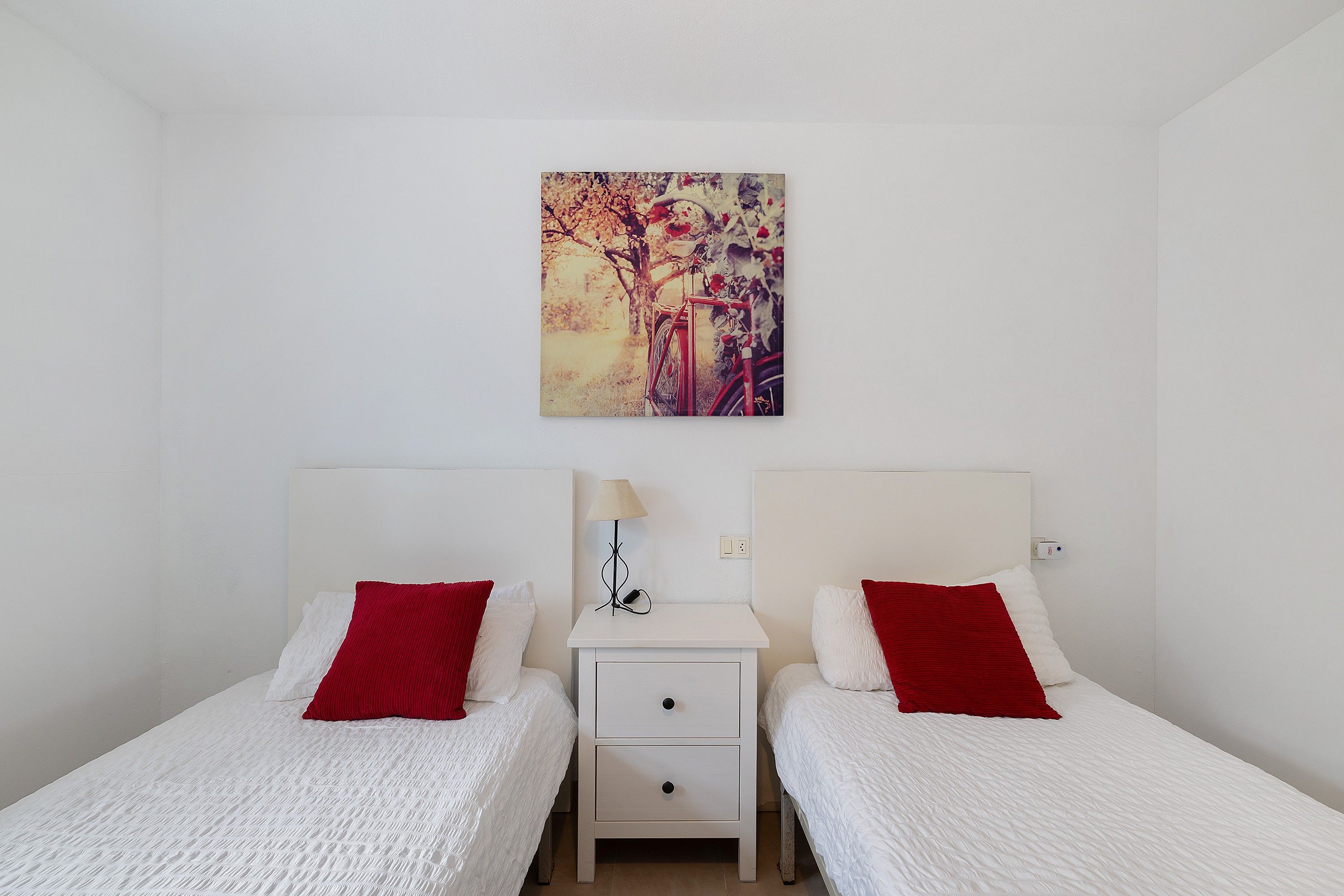 Un dormitorio con dos camas, cada una con una almohada roja, una mesita de noche con una lámpara y un cuadro en la pared.