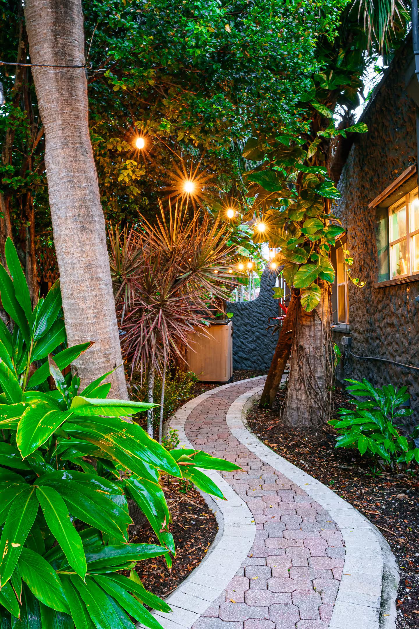 Un chemin pavé mène à un bâtiment à la façade en pierre, entouré d’une végétation luxuriante et illuminé par un éclairage décoratif.