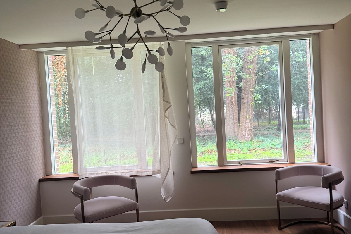 A bedroom with a large bed, two chairs, a modern chandelier, and large windows with a view of trees.