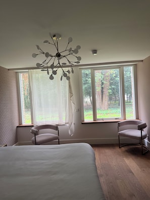 A bedroom with a large bed, two chairs, a modern chandelier, and large windows with a view of trees. - Parkhotel & Wellness Madeleine (Bavel)