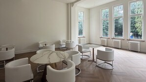 Une pièce spacieuse avec un plafond haut, de grandes fenêtres et différents types de sièges, notamment des tables rondes et des chaises.