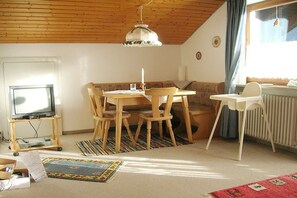 A cozy living room with a wooden ceiling, a television on a stand, a dining table with chairs, a sofa, a radiator, and a window with curtains. - Holiday apartment with 2 bedrooms (Oberstdorf)