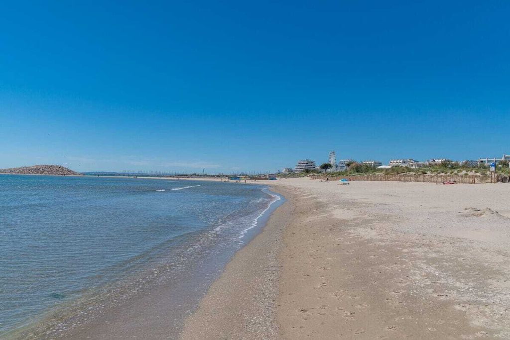 A beach with a clear sky, calm sea, and buildings in the distance.