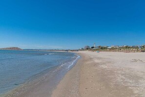 A beach with a clear sky, calm sea, and buildings in the distance.